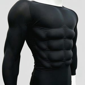 May include: Black compression shirt with padded muscle definition on the chest and abdomen. The long-sleeved shirt is designed to simulate a muscular physique. The garment is displayed on a white mannequin.