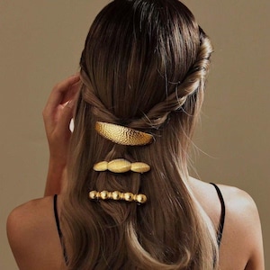 May include: Three gold-colored hair clips are displayed in a woman's hair. The top clip is a textured barrette, the middle clip is oval-shaped, and the bottom clip is a row of small beads. The woman's hair is light brown and styled in a twist.