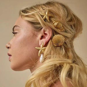 Könnte beinhalten: Haarschmuck und Ohrringe in Goldoptik mit Muscheln und Seesternen. Die Ohrringe haben ein Seestern-Design mit einer hängenden Perle. Der Haarschmuck umfasst eine Muschel und zwei Seestern-Haarspangen. Der Schmuck ist vor blondem Haar platziert.