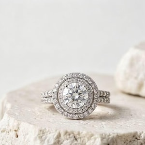 Puede incluir: Un anillo de compromiso plateado con una piedra central grande, redonda y transparente, rodeada por un doble halo de piedras más pequeñas. La banda también presenta piedras más pequeñas. El anillo se muestra sobre una superficie texturizada blanquecina.