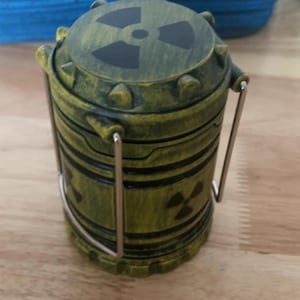 Pode incluir: Um recipiente cilíndrico com um acabamento verde e amarelo envelhecido, apresentando símbolos de radiação pretos. Possui uma tampa articulada e uma alça de metal. O recipiente é provavelmente um item decorativo ou um objeto de armazenamento.