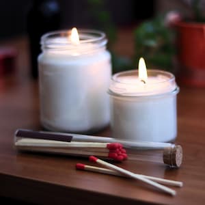 two lit candles in glass jars, along with a collection of red matchsticks, all placed on a wooden surface.