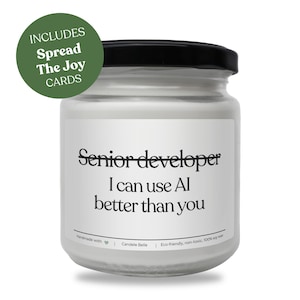 May include: A white candle in a clear glass jar with a black lid. The label features the text "Senior developer I can use AI better than you" in black font. A green circular badge in the upper left corner displays the text "INCLUDES Spread The Joy CARDS".