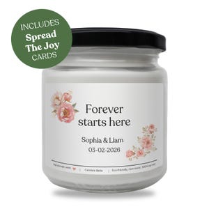 Puede incluir: Un tarro de vela blanco con tapa negra, con el texto "Forever starts here" y los nombres Sophia & Liam, con una fecha. El tarro está decorado con detalles florales e incluye un círculo verde que dice "Spread The Joy Cards".