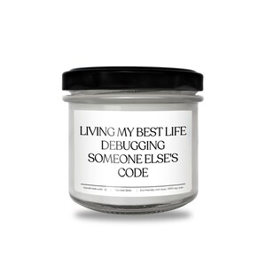 Peut inclure: Une bougie en pot de verre transparent avec un couvercle noir et une &eacute;tiquette blanche portant le texte noir : LIVING MY BEST LIFE DEBUGGING SOMEONE ELSE'S CODE. L'&eacute;tiquette inclut aussi : Handmade with heart | Candele Belle | Eco-friendly, non-toxic, 100% soy wax.