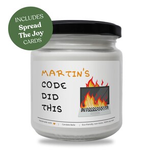 May include: A glass jar candle with a black lid and a white label featuring the text "MARTIN'S CODE DID THIS" in orange and black, alongside a graphic of a laptop engulfed in flames. A green circular badge reads "INCLUDES Spread The Joy CARDS".