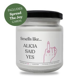 Könnte beinhalten: Weiße Sojawachskerze in einem Glas mit schwarzem Deckel. Auf dem Etikett steht "Smells like... ALICIA SAID YES" mit einer rosa Illustration einer Hand mit Ring. Enthält "Spread The Joy Cards".