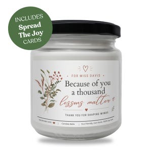 Può includere: Una candela bianca in barattolo con coperchio nero e un'etichetta che recita "Because of you a thousand lessons matter". L'etichetta include anche illustrazioni floreali e il testo "Spread The Joy Cards."