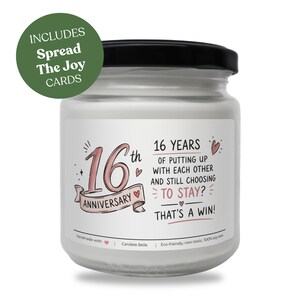 Puede incluir: Una vela blanca en un frasco de vidrio transparente con tapa negra. La etiqueta tiene texto en rosa y negro que dice 16th ANNIVERSARY y 16 YEARS OF PUTTING UP WITH EACH OTHER AND STILL CHOOSING TO STAY? THAT'S A WIN! Un círculo verde indica INCLUDES Spread The Joy CARDS.
