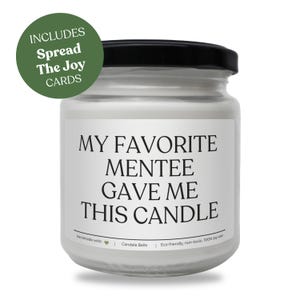 Può includere: Una candela in cera di soia bianca in un barattolo di vetro trasparente con coperchio nero. L'etichetta riporta il testo nero: MY FAVORITE MENTEE GAVE ME THIS CANDLE. Un badge circolare verde in alto a sinistra recita: INCLUDES Spread The Joy CARDS.