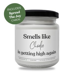 May include: A clear glass candle jar with a black lid and a white label featuring the black text Smells like Charlie is getting high again. A green circular badge in the upper left corner reads INCLUDES Spread The Joy CARDS in white text.