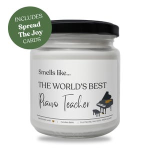 Può includere: Una candela bianca in un barattolo di vetro con coperchio nero. L'etichetta recita "THE WORLD'S BEST Piano Teacher" con un'illustrazione di pianoforte. Un cerchio verde indica "Spread The Joy Cards".