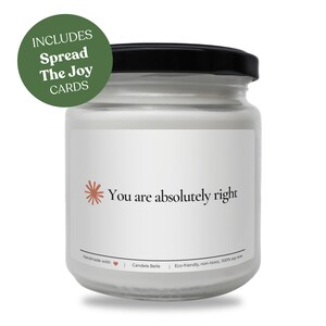 May include: A clear glass candle jar with a black lid and white wax. The label features a small coral-coloured starburst icon, the text "You are absolutely right," and a green circular badge stating "INCLUDES Spread The Joy CARDS."