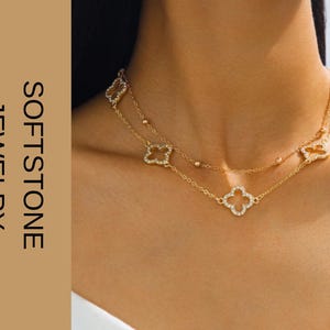 Può includere: Una collana dorata a due fili con pendenti a forma di trifoglio e piccole perline rotonde. La collana è indossata intorno al collo. L'immagine include anche il testo "SOFTSTONE JEWELRY" in orientamento verticale.