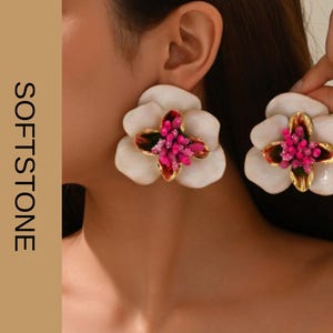 Puede incluir: Pendientes blancos con forma de flor con un centro dorado y un grupo de gemas rosas. Los pendientes son grandes y llamativos, con el texto "SOFTSTONE JEWELRY" en orientación vertical.