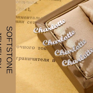 Puede incluir: Joyas de plata con el nombre "Charlotte" escrito en cursiva, expuestas en una caja de joyería. Las joyas se encuentran sobre un fondo beige. Las palabras "SOFTSTONE JEWELRY" están impresas verticalmente a la izquierda.