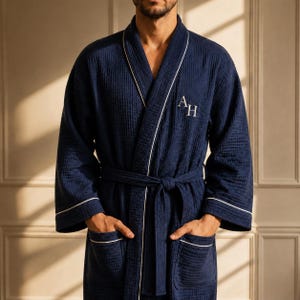 May include: A navy blue waffle-textured robe featuring white piping along the shawl collar, cuffs, and pockets. The robe includes a matching tie belt and is embroidered with the white capital letters AH on the left chest area of the garment.