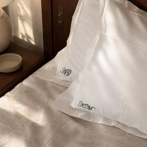 May include: Two white pillowcases with a flange border, each featuring black embroidered monogram initials. One pillowcase displays the letters AG, while the other shows the letters KM in a cursive script. The pillows rest on a light beige bedspread.