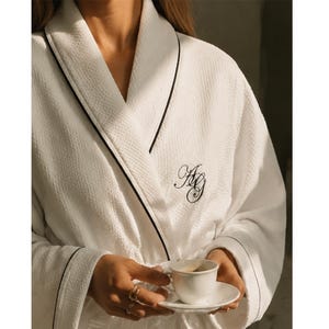 May include: A white waffle-textured robe featuring a black piped trim along the shawl collar and cuffs. The robe is embroidered with black cursive initials on the left chest. A small white ceramic espresso cup and saucer are held in front.