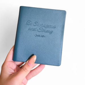 Könnte beinhalten: Ein teal-farbenes Notizbuch mit dem geprägten Text "Be Courageous and Strong - Josh 1:9." Das Cover hat eine strukturierte Oberfläche. Das Notizbuch wird in einer Hand vor weißem Hintergrund gehalten.