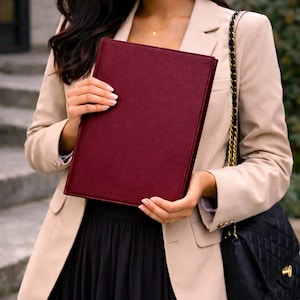 Puede incluir: Una carpeta portafolios rectangular de color burdeos intenso con un acabado mate y liso. La carpeta se sostiene en posición vertical, mostrando su color rojo oscuro sólido y sus bordes limpios y estructurados. Un organizador profesional.