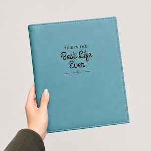 Könnte beinhalten: Ein blaugrünes Notizbuch mit einer glatten, matten Oberfläche. Auf dem Vorderdeckel befindet sich der schwarz aufgedruckte Text "THIS IS THE Best Life Ever", zentriert über einer kleinen Zierlinie mit einem Violinschlüssel in der Mitte.