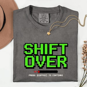 May include: A gray Comfort Colors t-shirt with the words "SHIFT OVER" in green pixelated font. Below the text is "HP" with a loading bar and the text "PRESS [COFFEE] TO CONTINUE". A gold necklace with a star pendant rests on the shirt.