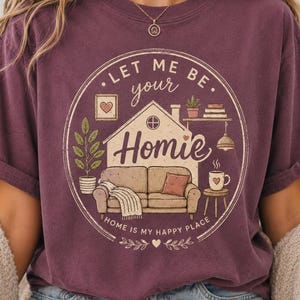 May include: Burgundy t-shirt with a circular graphic that reads "Let Me Be Your Homie." The graphic features a house, couch, and the phrase "Home is my happy place." The design includes a heart, plants, and a coffee cup.