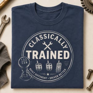 May include: Navy blue t-shirt with a white graphic. The graphic includes the text "CLASSICALLY TRAINED" in a circular design, along with a gear shift, pedals, and wrenches. The shirt also has the text "BUILT DIFFERENT • DRIVEN BETTER".