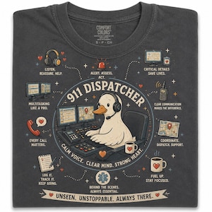 May include: Dark gray t-shirt featuring a cartoon duck wearing a headset, working at a 911 dispatcher station. The shirt includes the text "911 DISPATCHER" and other phrases like "Calm Voice, Clear Mind, Strong Heart" and "Unseen. Unstoppable. Always There."