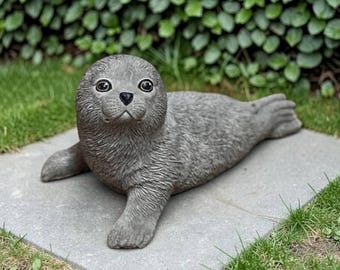 May include: A gray seal figurine with a textured surface, large dark eyes, and a small black nose. The seal is depicted in a resting position with its front flippers extended forward, showcasing detailed fur-like markings across its body.