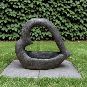 May include: A dark gray, textured, heart-shaped sculpture with a hollow center. The object features a smooth, curved top arching over a basin-like base, creating an open, abstract form with a weathered, stone-like appearance and finish.