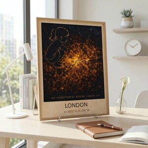 Op de afbeelding: Een ingelijste print met een kaart van Londen, verlicht met stadslichten. Het kunstwerk bevat een silhouet van een kind en de tekst "MY HOMETOWN, WHERE I GREW UP" en "LONDON". Coördinaten worden ook weergegeven.