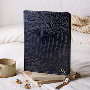 May include: A dark navy blue rectangular portfolio case with a textured crocodile skin pattern. The bottom right corner features the gold embossed initials SY. The case is standing upright on a stack of two books next to a ceramic mug and jewelry.