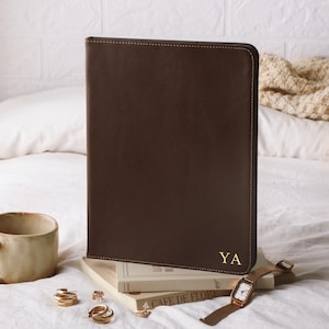 May include: A dark brown rectangular portfolio case with rounded corners and the gold embossed initials YA in the bottom right corner. The case is positioned upright on top of a stack of two books, next to a ceramic mug and a gold wristwatch.