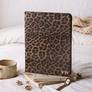 May include: A rectangular portfolio case featuring a brown and black leopard print pattern with the gold-colored initials YB embossed in the bottom right corner. The case stands upright on a stack of books next to a ceramic mug and a wristwatch.