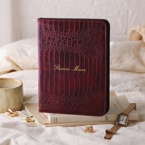 Pode incluir: Um diário de couro bordô com textura de crocodilo, com o nome "Sharon Moore" em letras douradas. O diário está sobre uma pilha de livros, ao lado de um relógio e anéis dourados. Uma caneca creme e um cobertor de malha estão no fundo.