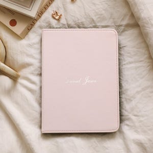 Puede incluir: Un diario de cuero rosa claro con el nombre "Daniel Jason" en escritura plateada. El diario está sobre una colcha blanca, junto a una taza de té, un reloj y libros.