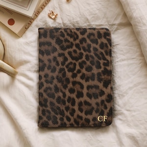 Può includere: Un portapassaporto con stampa leopardata marrone e nera con le iniziali dorate "CF". È appoggiato su una superficie bianca, insieme a una tazza di tè, un libro e un orologio.