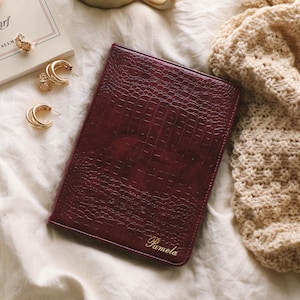 May include: A dark burgundy notebook with a textured crocodile skin pattern and the name Pamela embossed in gold script on the bottom right corner. The rectangular notebook is positioned on a white fabric surface next to gold hoop earrings.