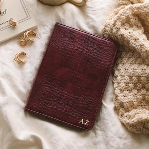 Könnte beinhalten: Ein weinrotes Lederjournal mit Krokodiltextur und goldenen Initialen "AZ" liegt auf einer weißen Oberfläche. Goldene Creolen, eine Uhr und ein Buch befinden sich in der Nähe. Ein beigefarbener Strickschal ist rechts drapiert.