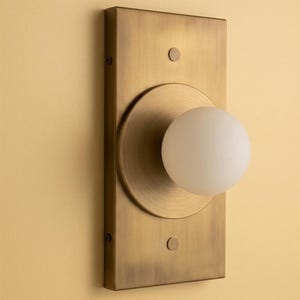 Puede incluir: Un aplique de pared con una placa rectangular de latón con acabado cepillado. Una placa circular de latón está centrada en el rectángulo, sosteniendo una pantalla esférica de vidrio blanco. Se observan dos pequeños tornillos circulares de latón.