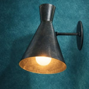 Puede incluir: Aplique de pared de metal oscuro con una pantalla cónica y luz cálida. La lámpara está montada sobre un brazo de metal oscuro y una base circular. El fondo es una pared turquesa texturizada. La pantalla tiene un diámetro de unos 20 cm.