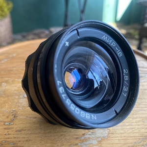 May include: A black camera lens with the text "МИР-1B 2.8/37" and the serial number "N86055737" printed on the side. The lens has a reflective glass surface and a ridged focus ring. The lens is resting on a wooden surface.