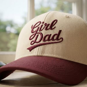 Może przedstawiać: Beżowa czapka z daszkiem z bordowym daszkiem. Czapka ma wyszyty napis "Girl Dad" bordową czcionką. Kapelusz jest wykonany z teksturowanej tkaniny, na rozmytym tle.