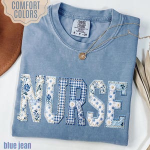 May include: A blue jean Comfort Colors t-shirt with the word "NURSE" appliqued across the front. The letters are a mix of floral, polka dot, and gingham patterns. A gold necklace with a small pendant rests on the shirt.