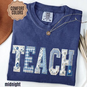 May include: A navy blue Comfort Colors t-shirt with the word "TEACH" appliqued in various blue floral and gingham patterns. A gold necklace with a pendant rests on the shirt. The shirt is labeled "midnight".