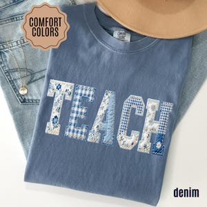 May include: A blue Comfort Colors t-shirt with the word "TEACH" embroidered in a patchwork design. The letters feature floral, gingham, and solid blue patterns. A tan hat and denim jeans are also visible.