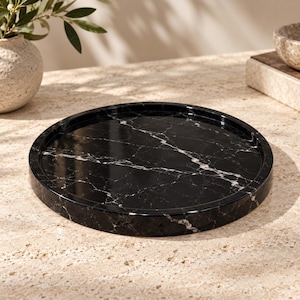 May include: A circular black marble tray featuring a polished finish with prominent white veining throughout the surface. The tray has a raised edge and a smooth, reflective texture, showcasing the natural stone pattern in a simple, round design.