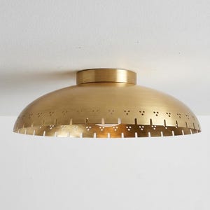 May include: A brass-colored, semi-spherical ceiling light fixture. The light has a perforated edge with small, evenly spaced holes. The fixture is mounted on a white ceiling, creating a contrast with the warm metallic finish.